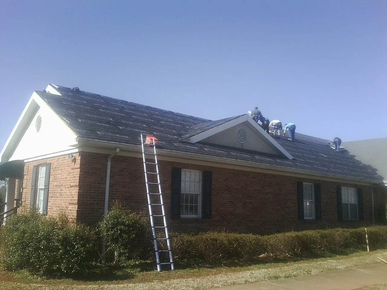 Crew tearing shingles off a brick ranch roof with a ladder set — Emergency Roof Repair work by our crew in Baldwyn
