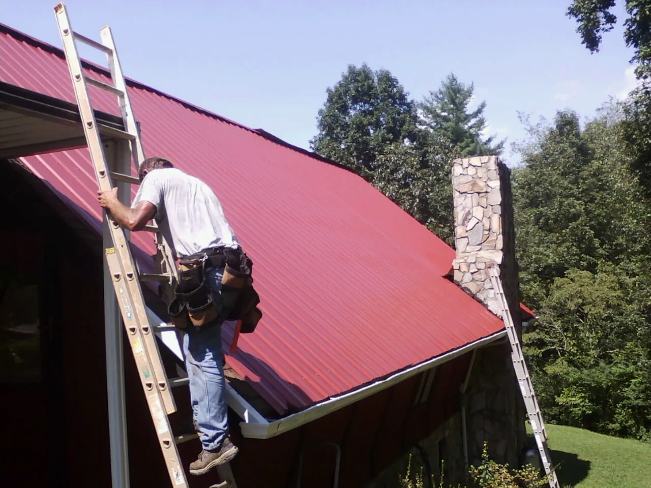 Roofer on a ladder beside a red metal roof and stone chimney — Soffit & Fascia Repair by Roofing Brothers in Baldwyn